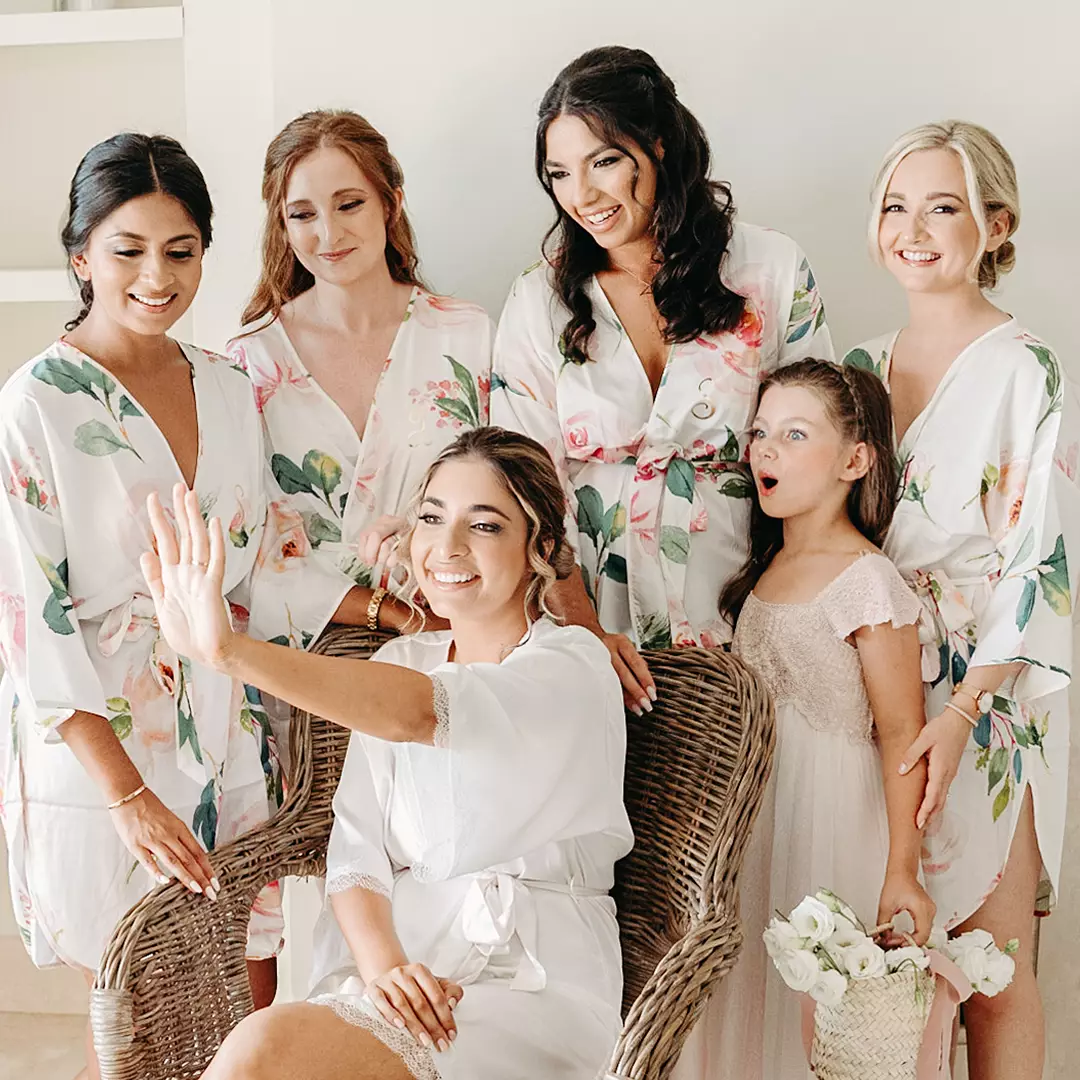 A bride shows her ring while sitting in a chair, surrounded by bridesmaids and a flower girl, all wearing floral robes and smiling.
