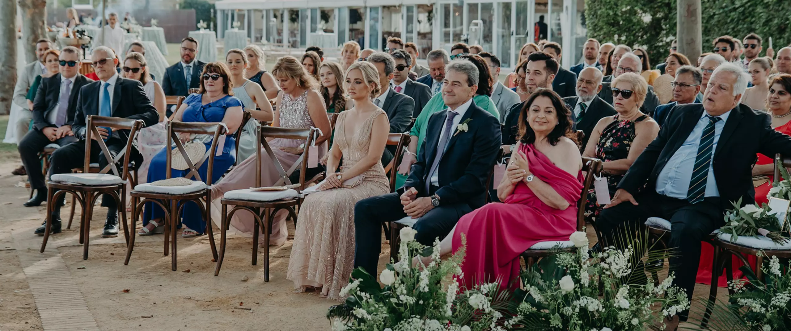 A group of formally dressed guests sit in rows of chairs at an outdoor wedding ceremony, with greenery and flowers in the foreground.