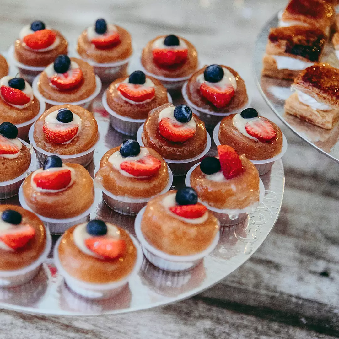 A tray of small pastries topped with cream, sliced strawberries, and blueberries sits next to a plate of layered cream-filled desserts on a wooden surface.