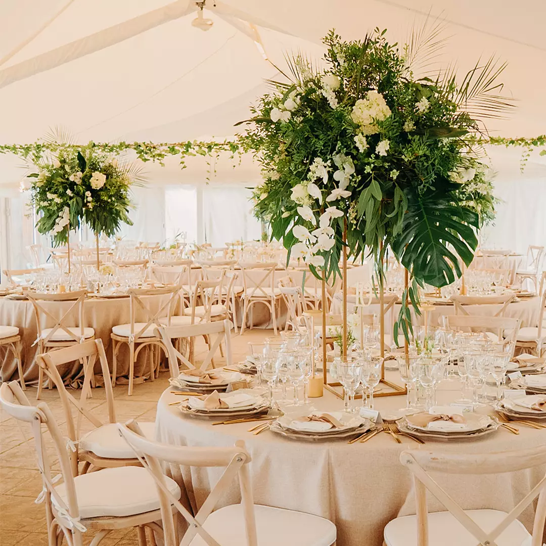 Elegant banquet tables set with plates, glasses, and cutlery, featuring tall green and white floral centerpieces in a bright, decorated event tent.