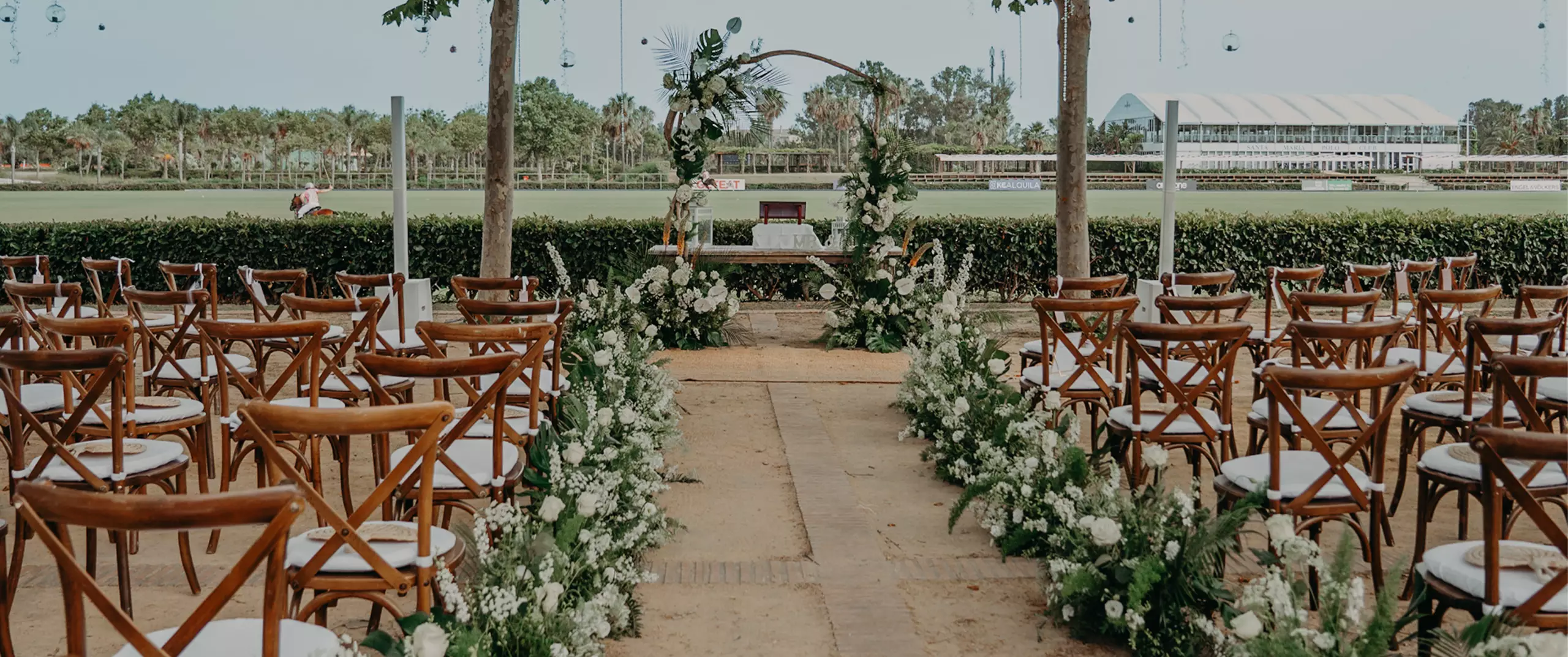 Outdoor wedding ceremony setup with wooden chairs in rows, floral arrangements lining the aisle, and a decorated arch in the background.