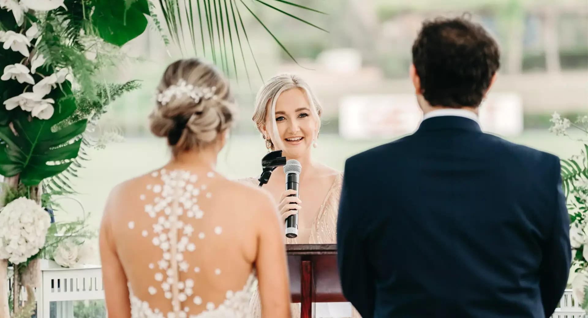 A woman officiates a wedding ceremony, speaking into a microphone to a bride and groom standing side by side.