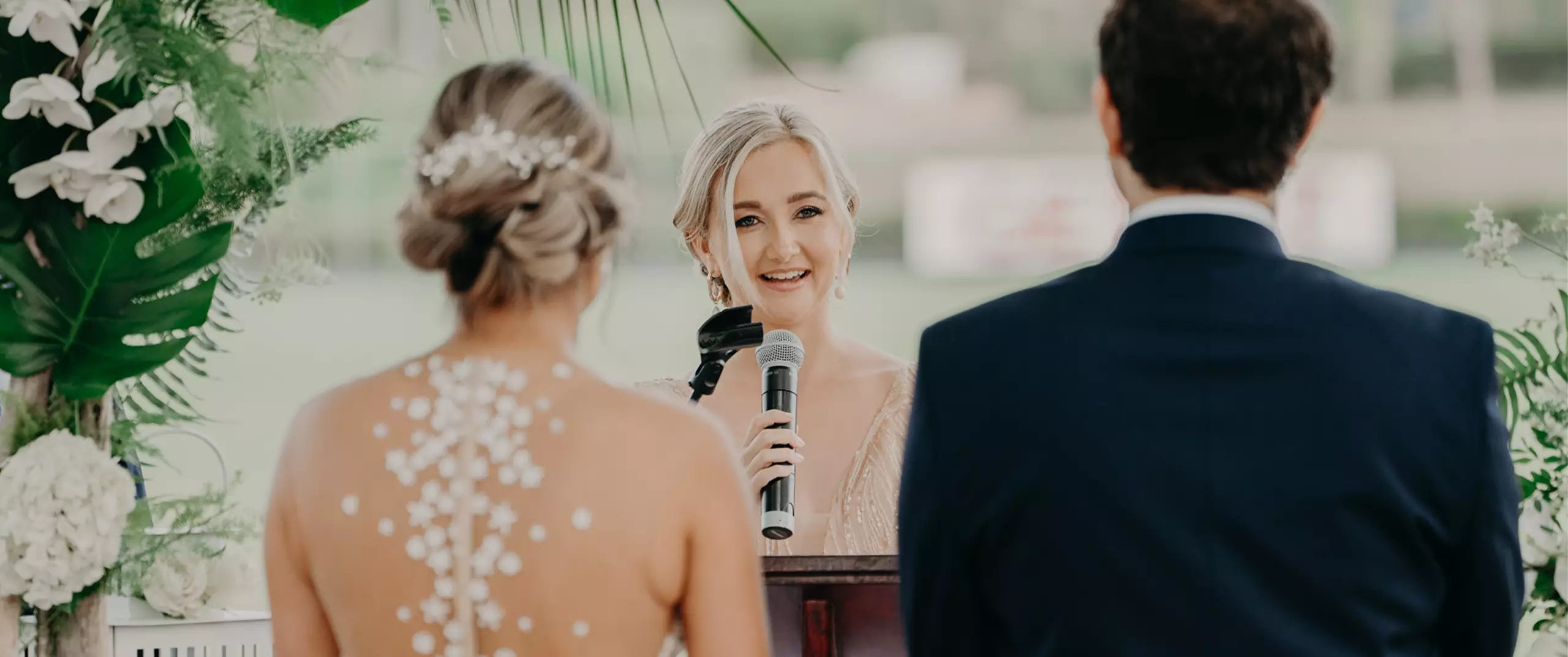 A woman speaks into a microphone at a wedding ceremony, standing at a podium between a bride in a beaded dress and a groom in a dark suit.