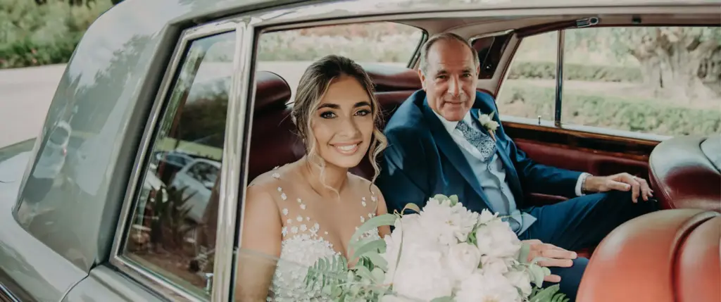 A bride in a white dress sits beside an older man in a suit inside a car, both smiling at the camera.