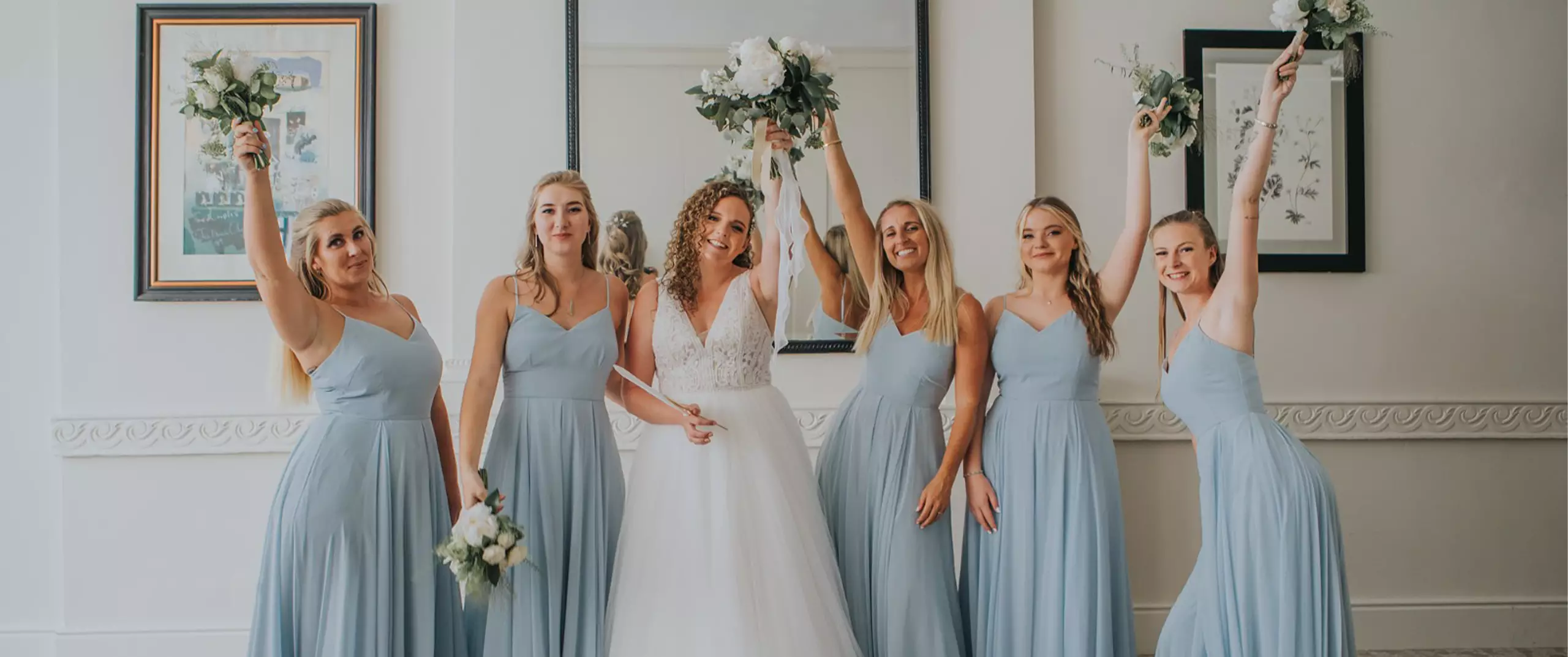 A bride in a white gown stands with five bridesmaids in matching light blue dresses, all holding bouquets and posing in front of a large mirror indoors.