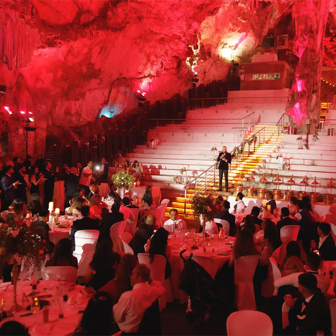 A formal event with seated guests in a cave-like venue, featuring red lighting and a staircase where a person stands and speaks.