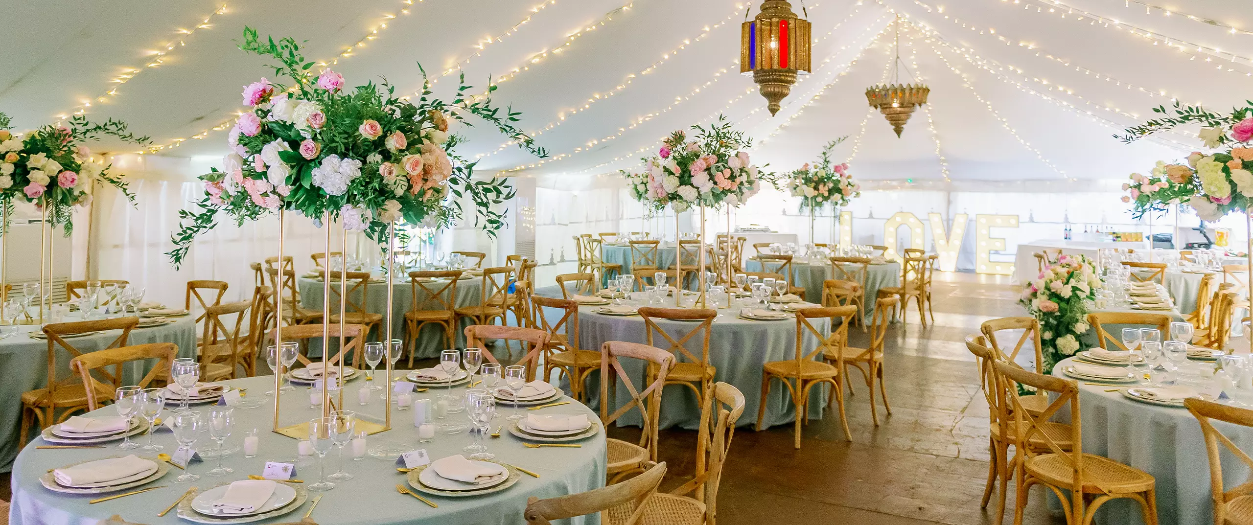 A decorated wedding reception tent with round tables, floral centerpieces, wooden chairs, string lights, and large illuminated 