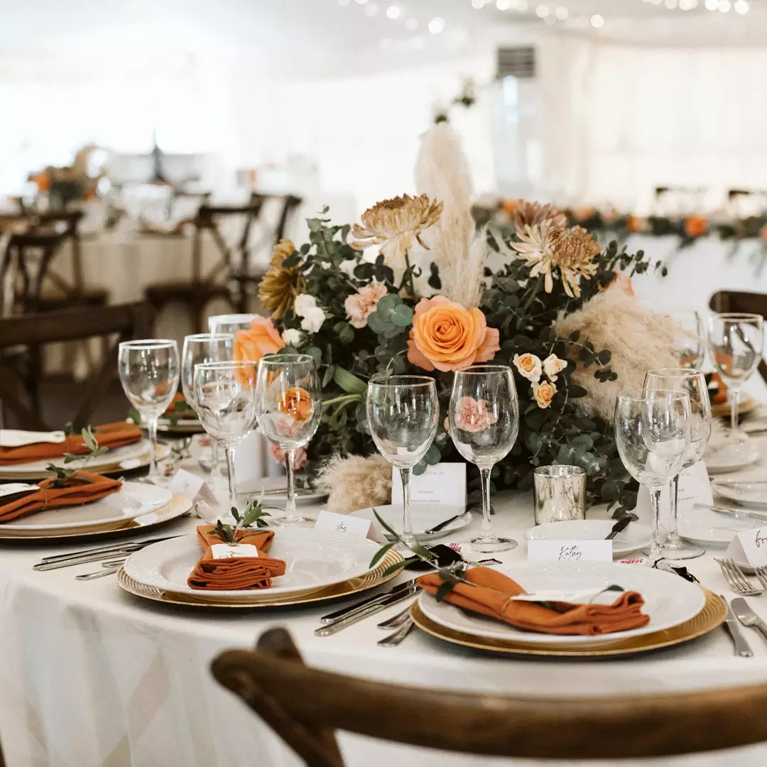 A round banquet table set with plates, cutlery, wine glasses, orange napkins, place cards, and a floral centerpiece with orange and white flowers.