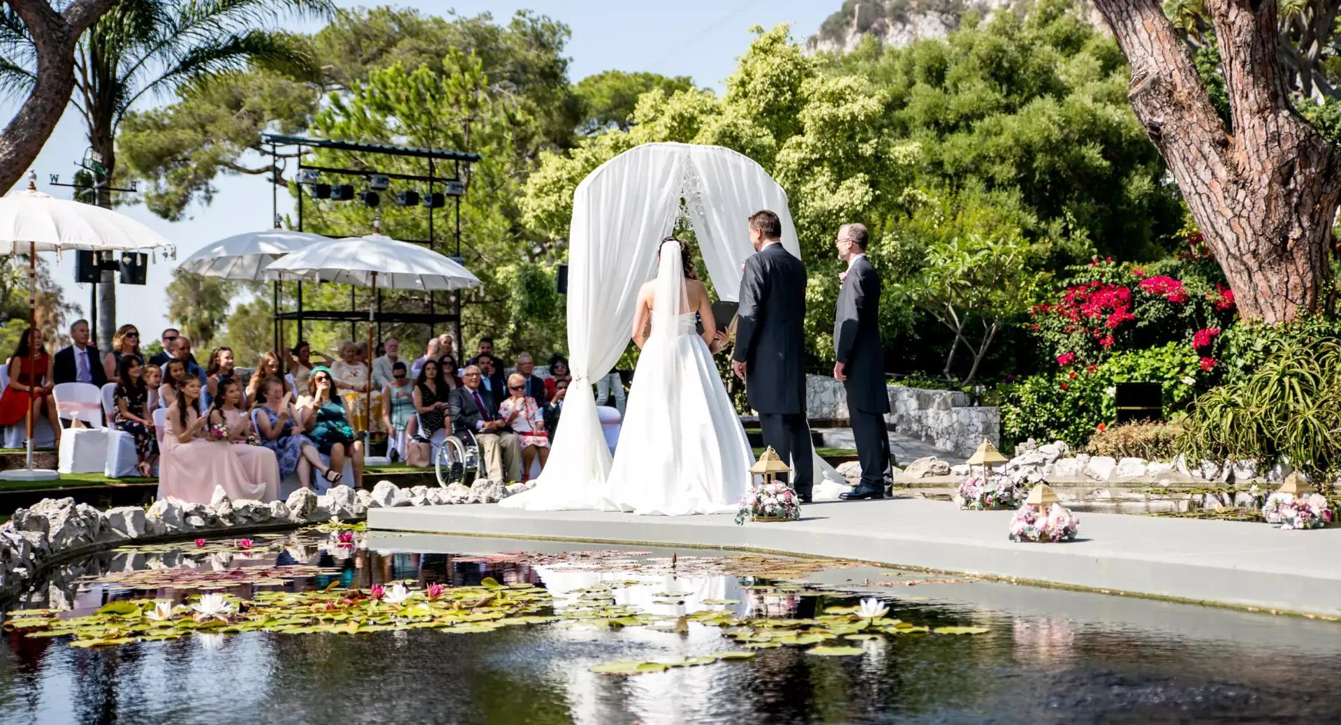 A bride and groom stand with an officiant under a white arch during an outdoor wedding ceremony, with guests seated nearby and a pond in the foreground.