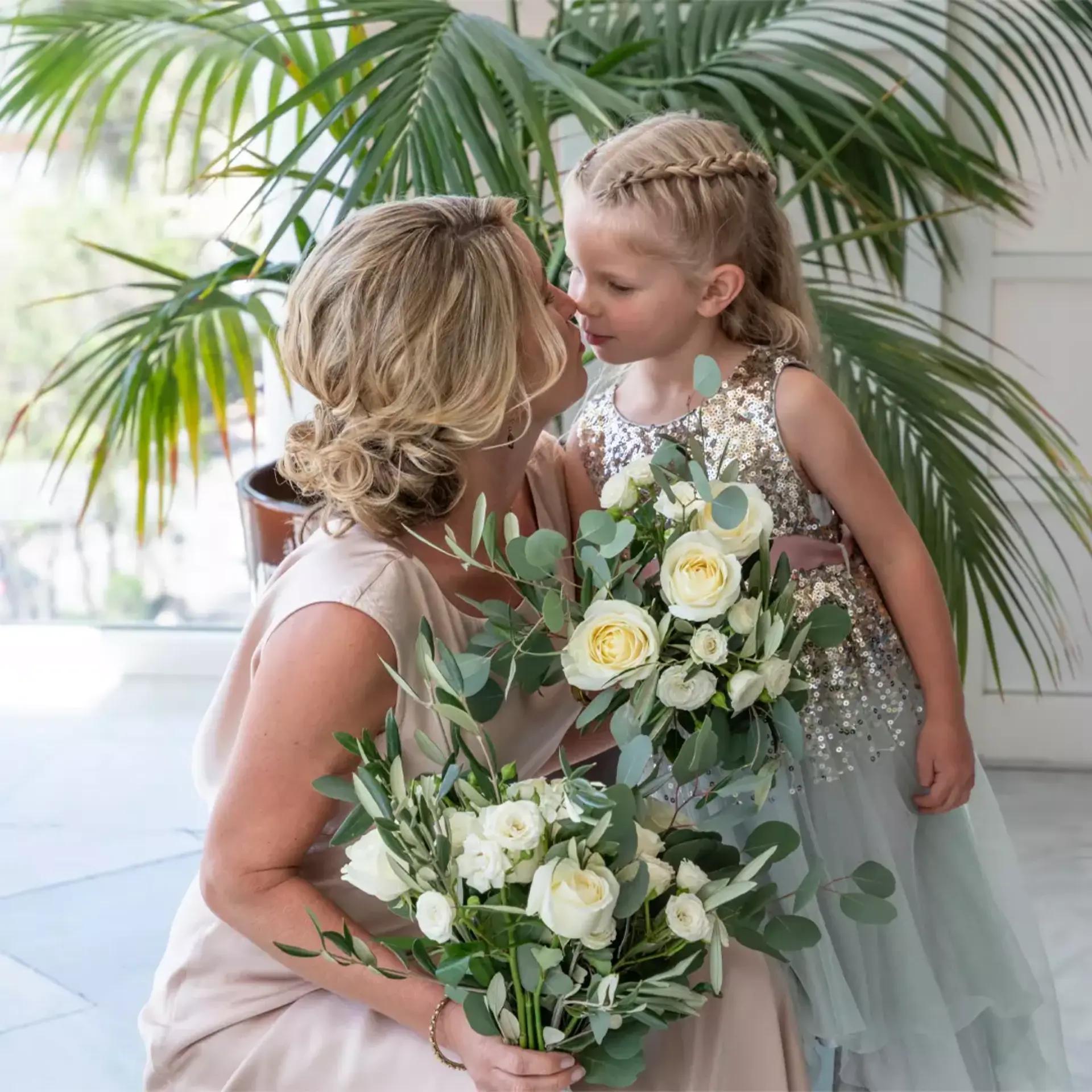 An adult and a child in formal attire share a close moment while holding bouquets of white flowers and greenery, with a large plant in the background.