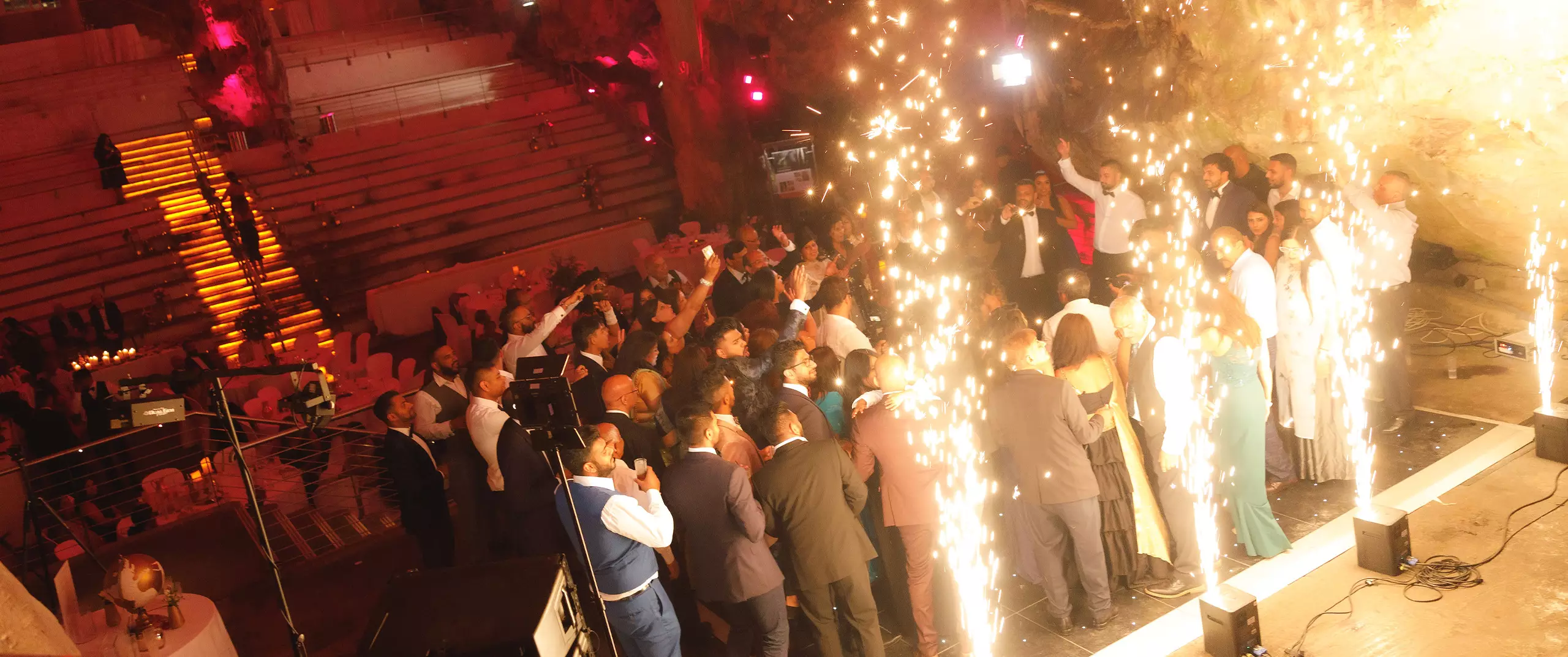 A large group of people dressed formally gather on a stage with bright sparks and lights, while others are seated in the background of an indoor venue.