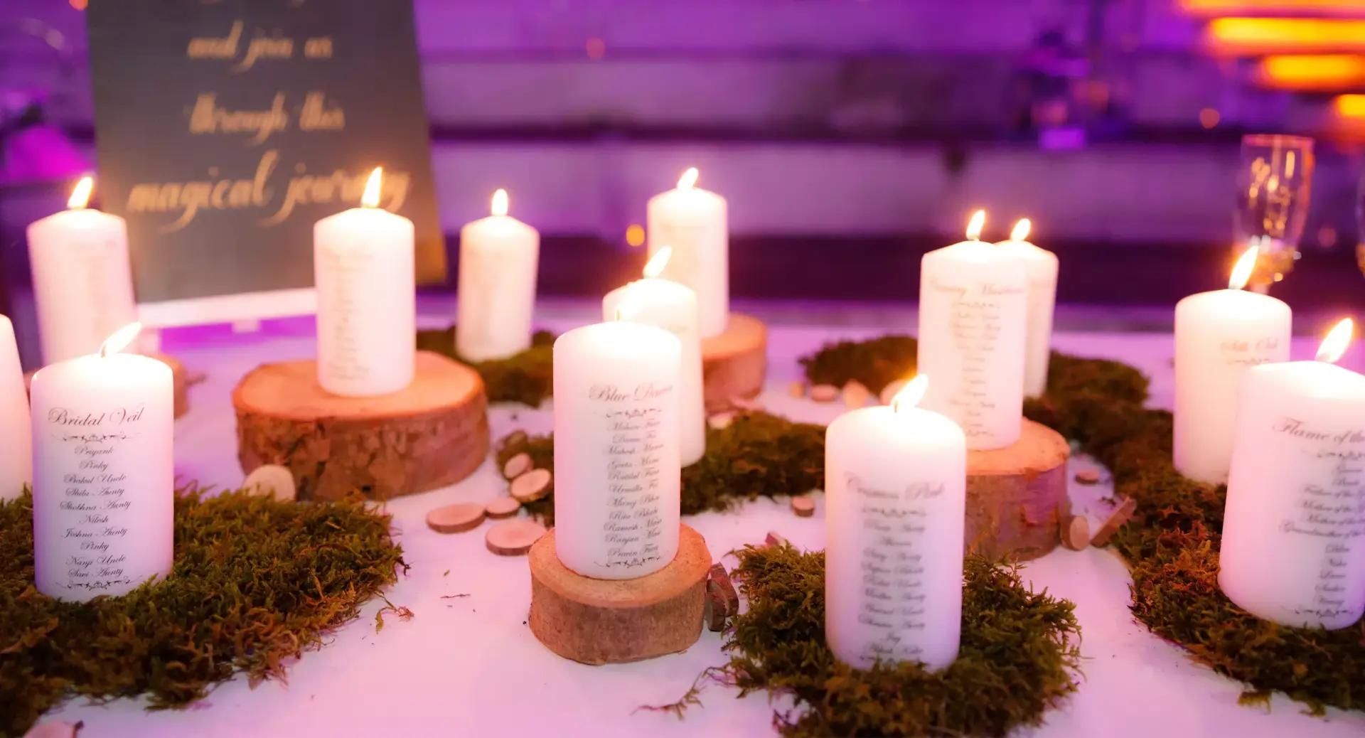 Multiple lit white candles with printed text stand on wooden slices and moss, arranged on a table with a purple-lit background and a blurred sign in the distance.
