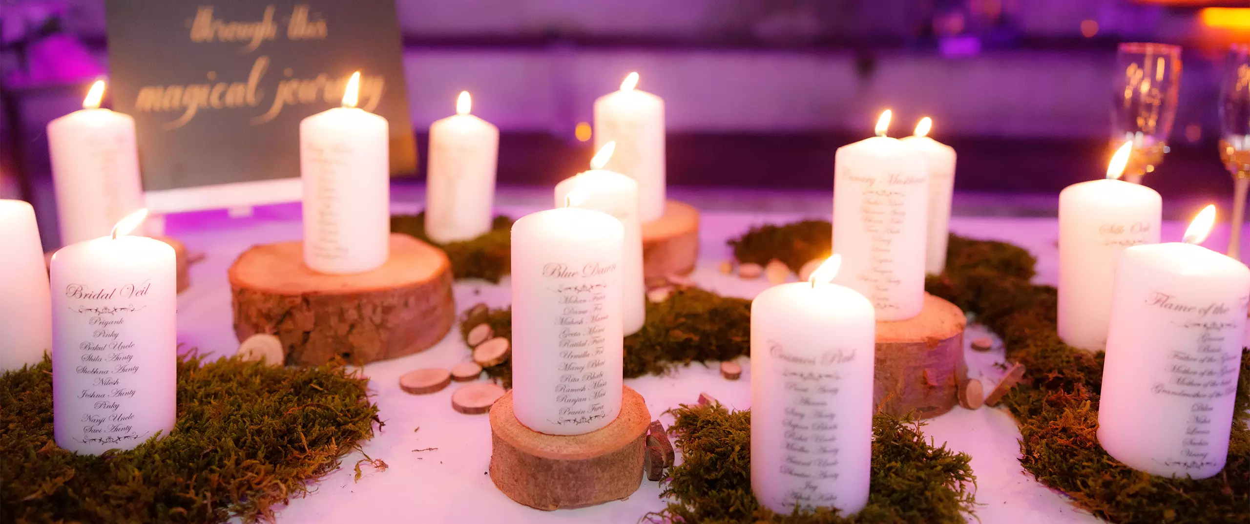 Several lit white candles with printed text are arranged on wood slices and moss, creating a centerpiece on a table with a blurred sign and purple lighting in the background.