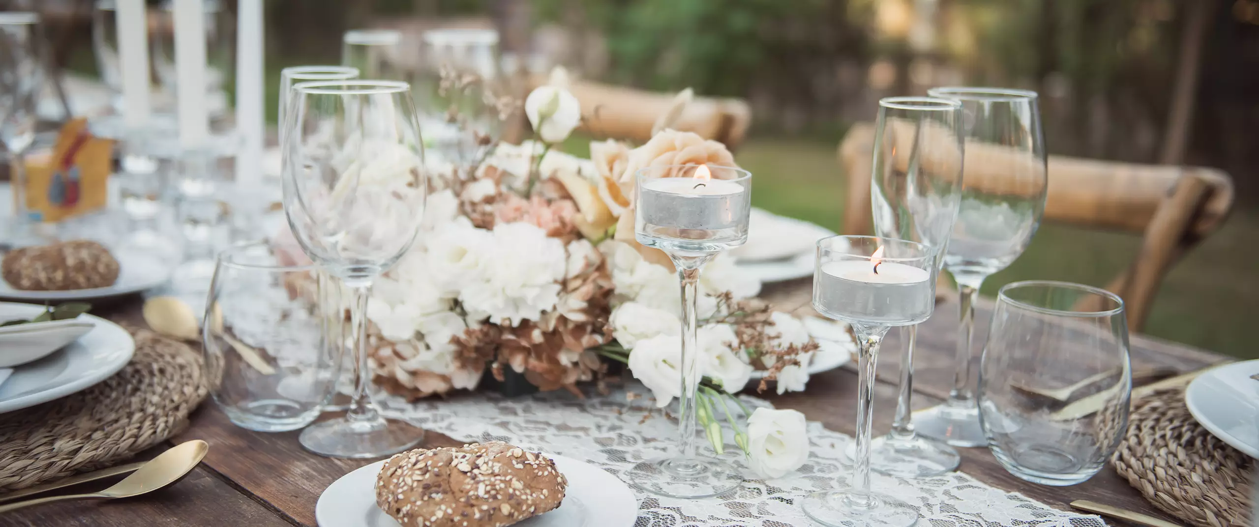 Elegant outdoor table setting with glassware, candles, white and peach floral centerpiece, plates, and seeded bread rolls on a rustic wooden table with woven placemats.