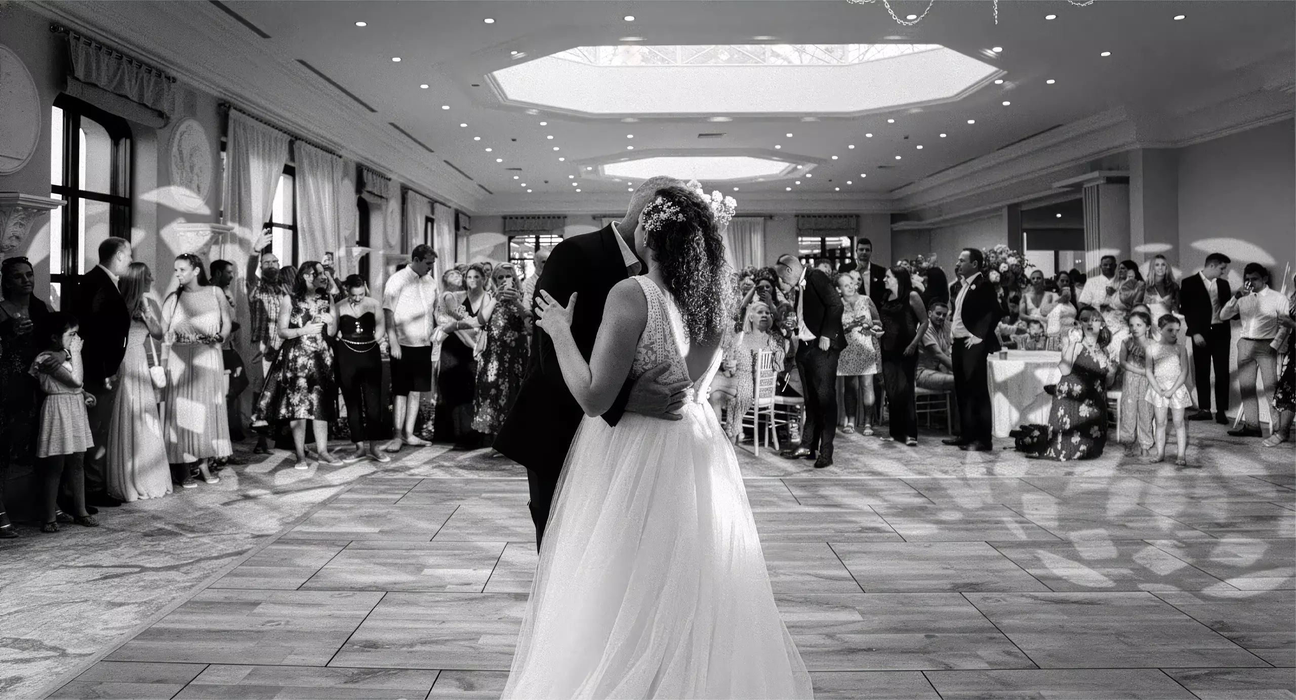 A bride and groom share their first dance in a large, elegant ballroom as wedding guests watch and take photos.