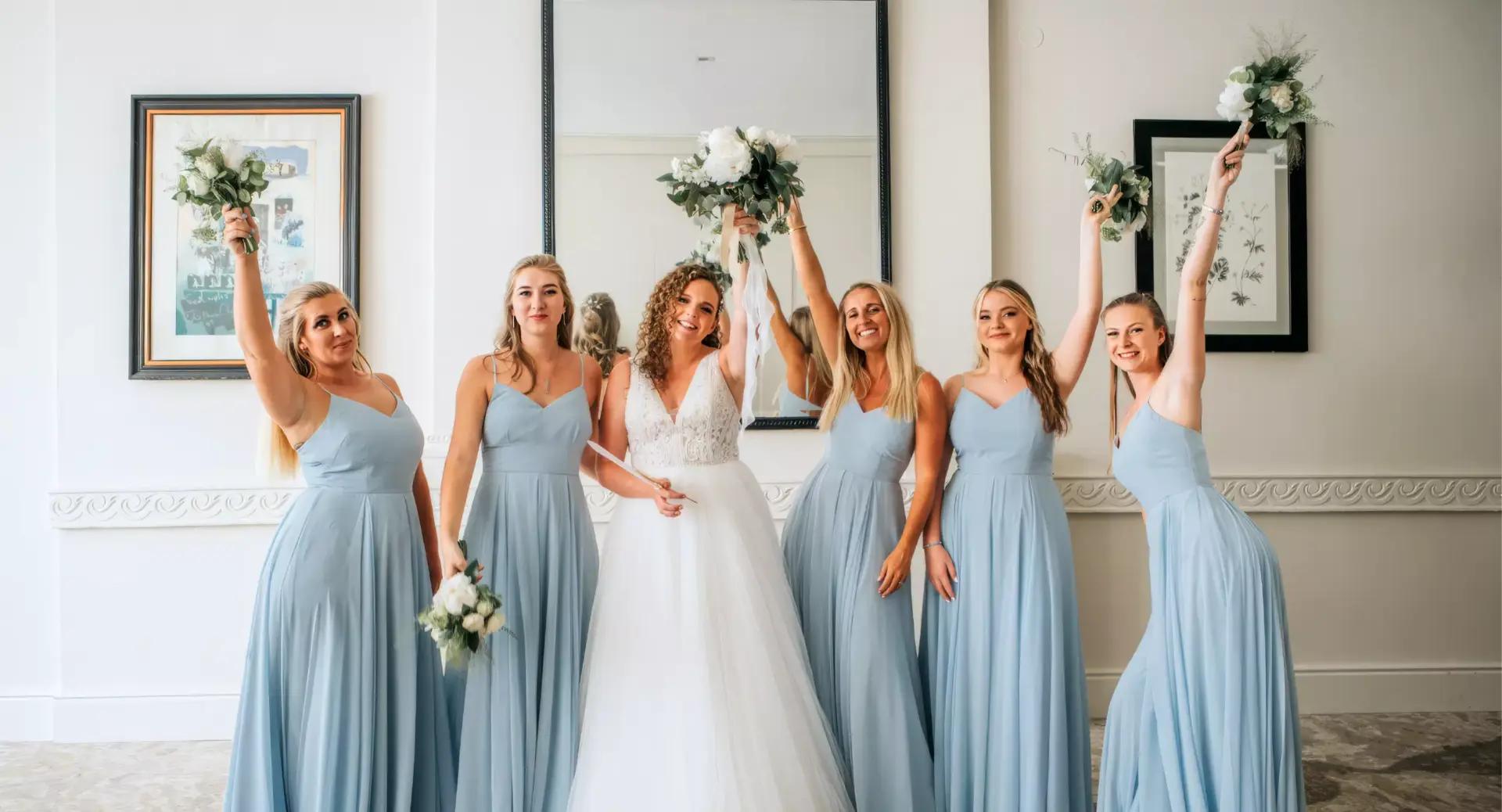 A bride in a white gown stands with four bridesmaids in matching light blue dresses, all holding bouquets and smiling in a brightly lit room.