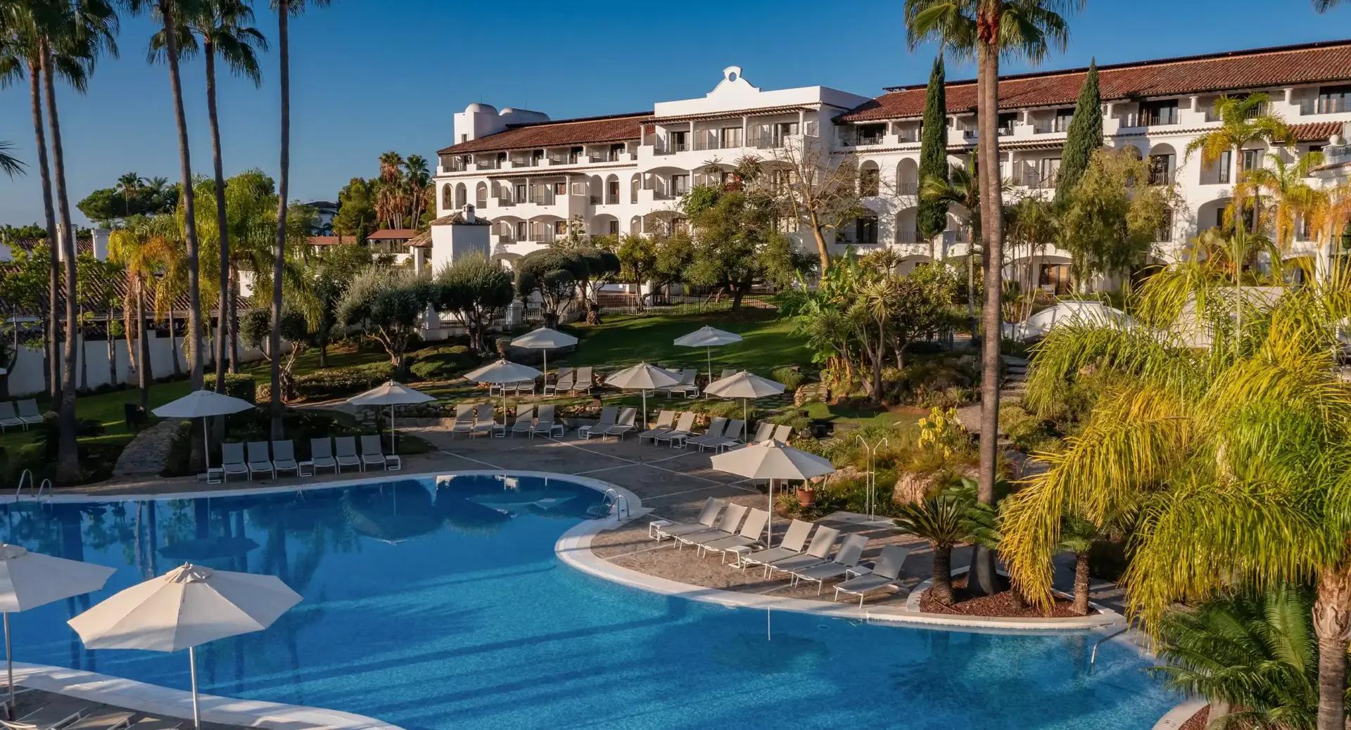 A large hotel with white walls overlooks a landscaped pool area with lounge chairs, umbrellas, palm trees, and gardens under a clear blue sky.